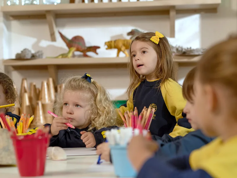 Crianças da Educação Infantil no ateliê, com lápis de cor na mesa; uma delas fala com a turma